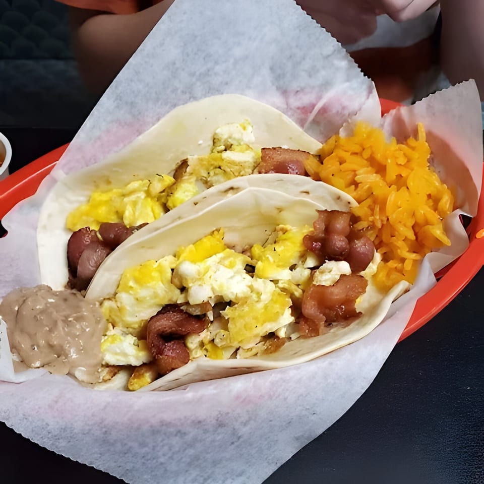 Two breakfast tacos with scrambled eggs, bacon, refried beans, and orange rice in a red basket.