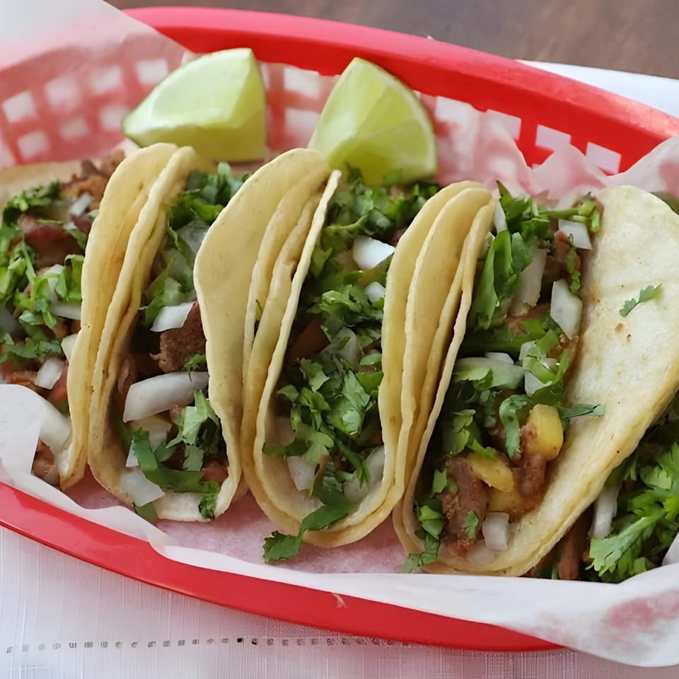 Three La Familia Cafe Mexican street tacos with meat, cilantro, onions, and lime wedges in a red basket.